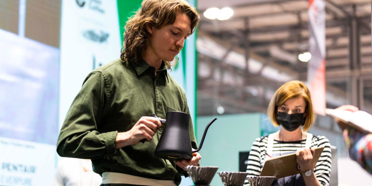 Ein Barista gießt mit einer Kaffeekanne beim Brühen von Kaffee auf einer Messe, während eine Frau mit Maske im Hintergrund Notizen macht.