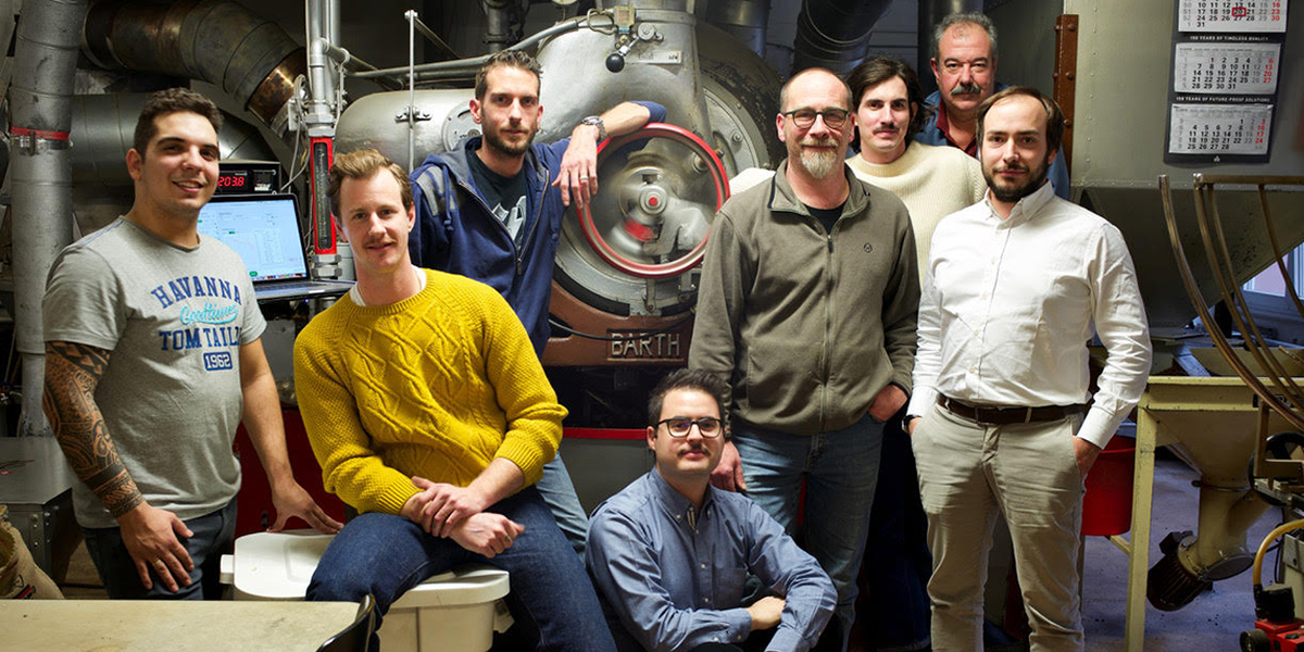 Eine Gruppe von sieben Männern in einer Kaffeerösterei vor einer großen Röstmaschine.