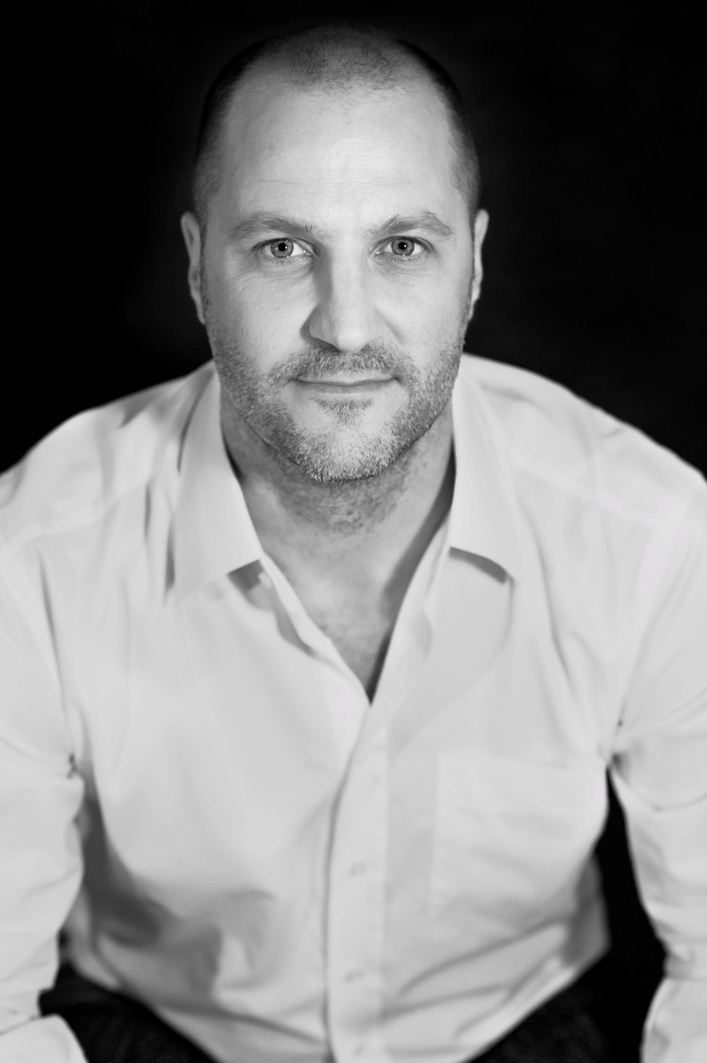 Porträt eines Mannes in Schwarz-Weiß Schwarz-Weiß-Fotografie eines Mannes in einem weißen Hemd, der in die Kamera schaut und vor einem dunklen Hintergrund sitzt.