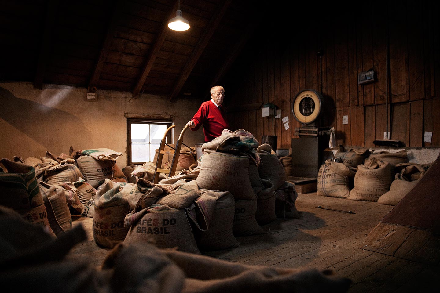 Ein älterer Mann steht in einem rustikalen Lagerhaus voller Kaffeesäcke aus Brasilien, umgeben von Holzverkleidung und warmer Beleuchtung.